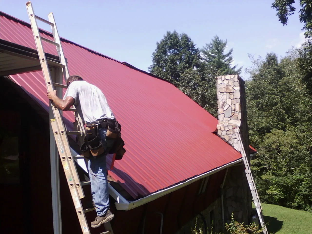 Roofer on a ladder beside a red metal roof and stone chimney — Soffit & Fascia Repair by Roofing Brothers in Amanda
